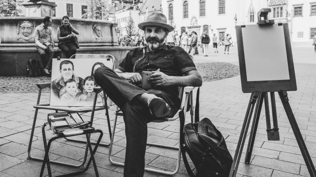 Grayscale Photo of Man Wearing Black Dress Shirt With Hat Sitting on Folding Armchair Beside Family Sketch